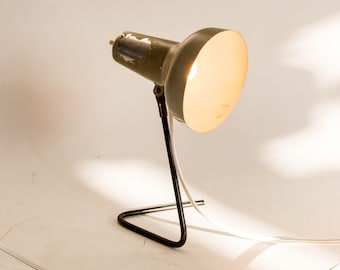May include: A vintage desk lamp with a green metal shade and a black metal base. The lamp has a white cord and is turned on, illuminating the room.