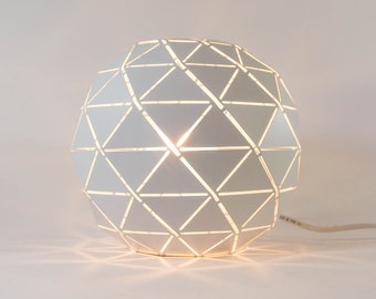 Könnte beinhalten: Eine weiße geometrische Tischlampe mit einem dreieckigen Muster. Die Lampe ist eingeschaltet und strahlt ein warmes Licht aus.