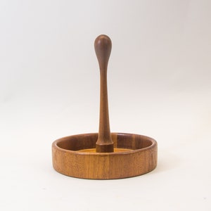 Puede incluir: Un porta anillos de madera con una base redonda y una perilla alta, delgada y redondeada. El soporte está hecho de madera marrón claro.