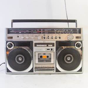 Puede incluir: Radiocasete Toshiba plateado y negro con una cinta de cassette insertada. La radio tiene un asa, una antena y dos altavoces grandes. El número de modelo es MT15. El radiocasete está sobre una superficie blanca.