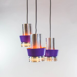 Op de afbeelding: Een moderne hanglamp met drie lampen, met een paarse kap en cilindrische zilveren kappen. Elke kap heeft een paarse basis en een warme oranje gloed. De lamp hangt aan zwarte snoeren.