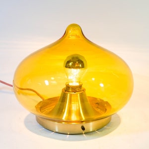 Peut inclure: Une lampe de table en verre jaune avec une base dorée et une ampoule à l'intérieur. La lampe a une forme arrondie en forme de larme.