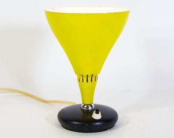 May include: A yellow table lamp with a black base and a round, cone-shaped shade. The lamp has a cord and a switch on the base.