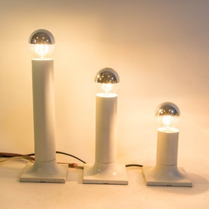 Może przedstawiać: Trzy białe lampy stołowe z srebrnymi żarówkami. Lampy są różnych rozmiarów i są ułożone w rzędzie od największej do najmniejszej.