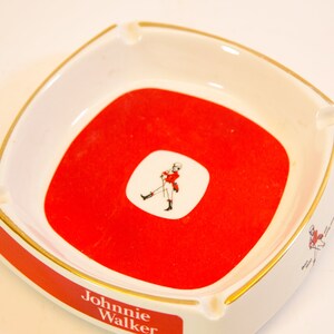 May include: Square white ashtray with a red center and gold trim. The center features a white square with a figure in red. The ashtray has the words "Johnnie Walker" in red on the side.