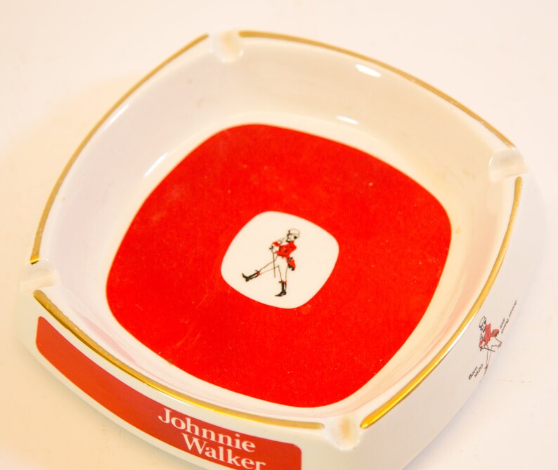 May include: Square white ashtray with a red center and gold trim. The center features a white square with a figure in red. The ashtray has the words "Johnnie Walker" in red on the side.