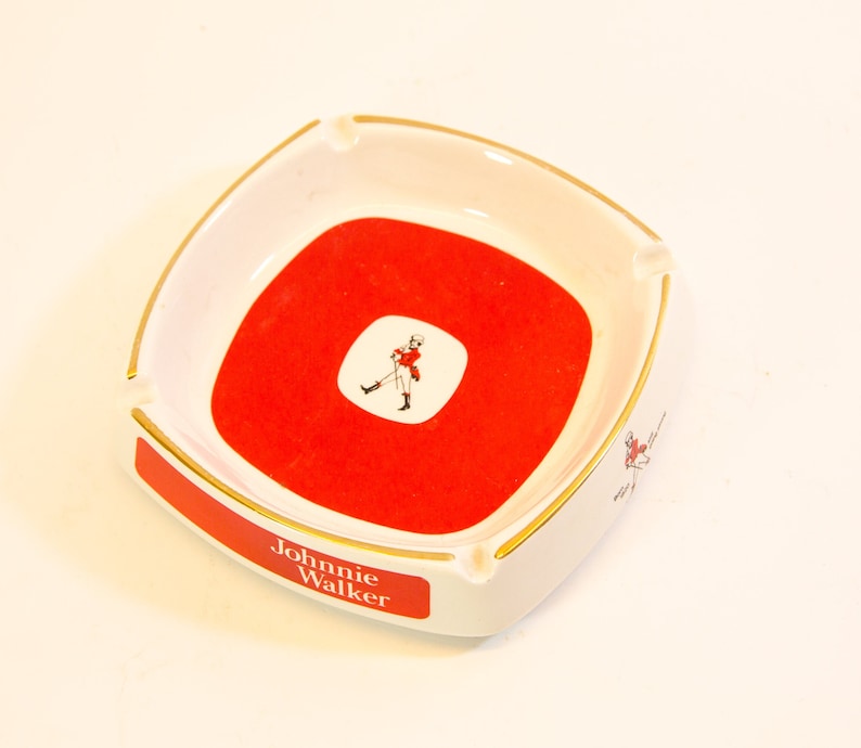 May include: Square white ashtray with a red center and gold trim. The ashtray features the Johnnie Walker logo and text. A small illustration of a man in a red coat is in the center. The ashtray has a curved design.