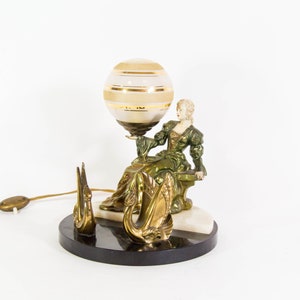 Puede incluir: Una l&aacute;mpara de mesa vintage con una figura de mujer verde y dorada sosteniendo una pantalla de l&aacute;mpara en forma de globo. La base de la l&aacute;mpara es de m&aacute;rmol negro y presenta dos figuras de cisnes dorados.