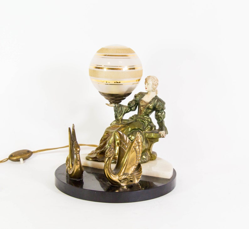 Puede incluir: Una l&aacute;mpara de mesa vintage con una figura de mujer verde y dorada sosteniendo una pantalla de l&aacute;mpara en forma de globo. La base de la l&aacute;mpara es de m&aacute;rmol negro y presenta dos figuras de cisnes dorados.