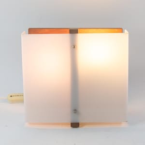 Puede incluir: Una lámpara de mesa blanca con una base de madera y un marco de metal. La lámpara tiene una pantalla de vidrio esmerilado y un cable con un interruptor.