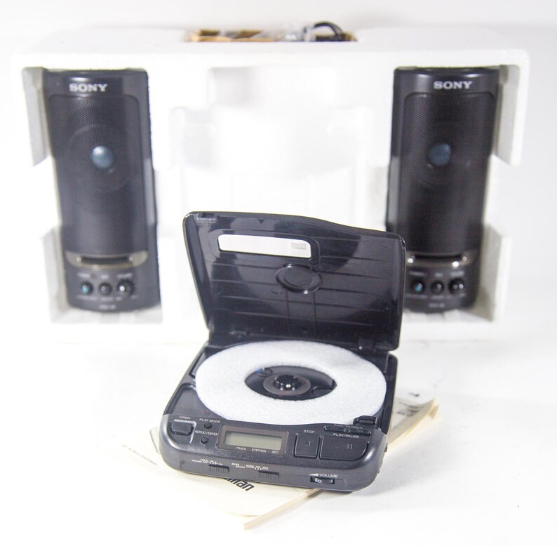 May include: A black Sony CD player with a white disc inside and two black Sony speakers on either side. The speakers are in white boxes with the word "SONY" printed on them.