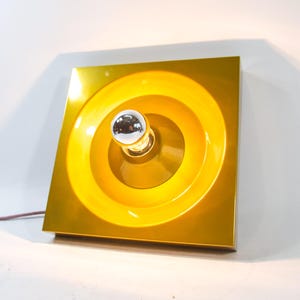 Op de afbeelding: Een vierkante, goudkleurige wandlamp met een cirkelvormig, verzonken ontwerp. Een verchroomde, bolvormige gloeilamp is gecentreerd in het ontwerp. De lamp heeft een glanzende afwerking en een bruin netsnoer.