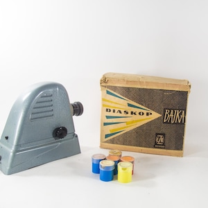 May include: A vintage slide projector with a gray body and a black lens. The projector is labeled "DIASKOP" and "BAIKA" on the box. The box is brown with yellow and blue stripes. There are six small colorful containers next to the projector.