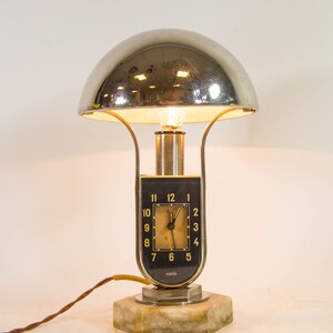 Peut inclure: Une lampe de table en chrome et en marbre avec un cadran d'horloge qui indique "norch". La lampe a un abat-jour en forme de d&ocirc;me et une base argent&eacute;e.