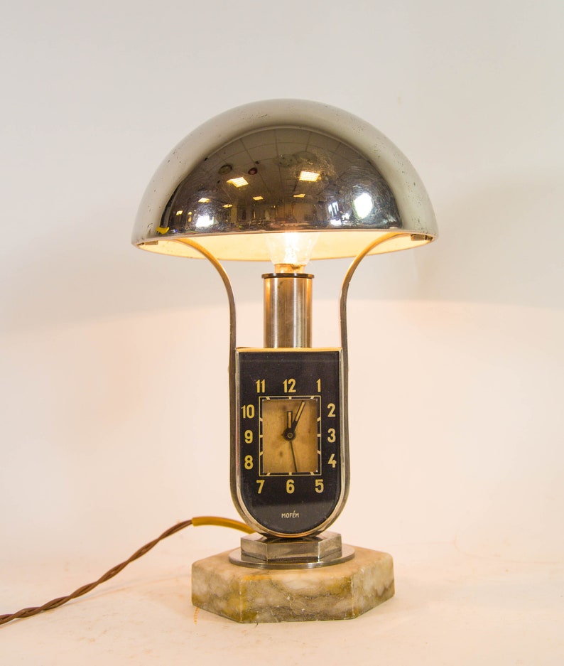 Peut inclure: Une lampe de table en chrome et en marbre avec un cadran d'horloge qui indique "norch". La lampe a un abat-jour en forme de d&ocirc;me et une base argent&eacute;e.