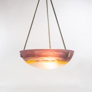Pu&ograve; includere: Una lampada a sospensione con un paralume a forma di ciotola. Il paralume presenta una miscela di colori, tra cui viola, arancione e giallo, con un interno bianco. La lampada &egrave; sospesa da tre aste metalliche.