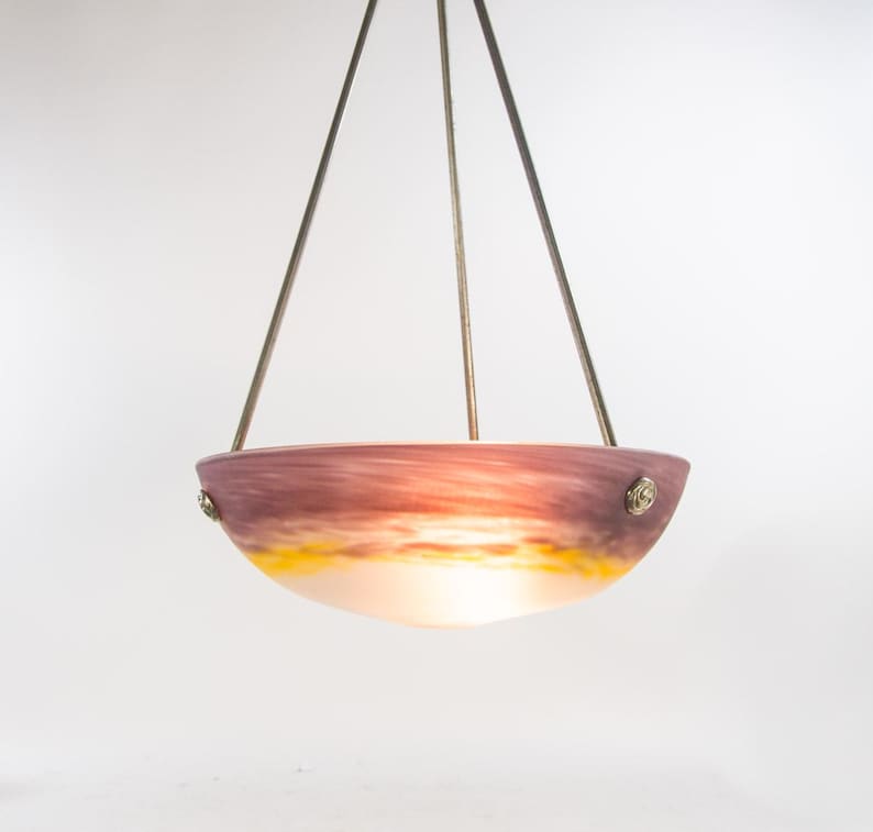 Pu&ograve; includere: Una lampada a sospensione con un paralume a forma di ciotola. Il paralume presenta una miscela di colori, tra cui viola, arancione e giallo, con un interno bianco. La lampada &egrave; sospesa da tre aste metalliche.