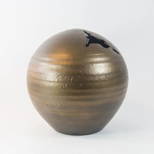 Könnte beinhalten: Eine runde, goldfarbene Keramikvase mit strukturierter Oberfläche und einer kleinen Öffnung oben.