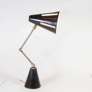 Może przedstawiać: Czarna lampa biurkowa z regulowanym srebrnym ramieniem i czarnym, stożkowym kloszem z wycięciami. Lampa stoi na białym tle.