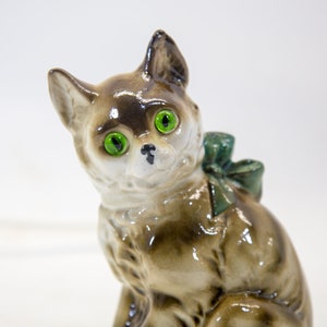 May include: A ceramic figurine of a brown and white cat with green eyes and a green bow. The cat is sitting with its head tilted to the side.