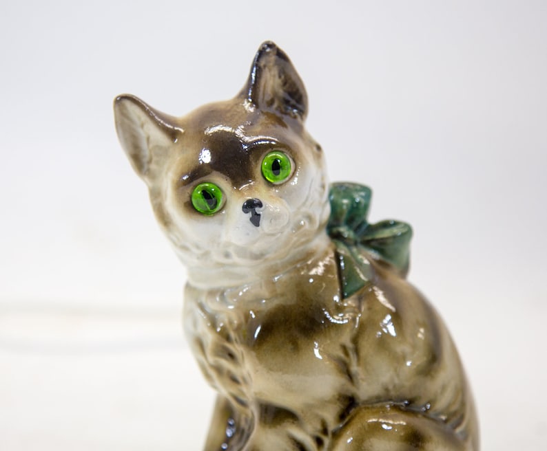 May include: A ceramic figurine of a brown and white cat with green eyes and a green bow. The cat is sitting with its head tilted to the side.