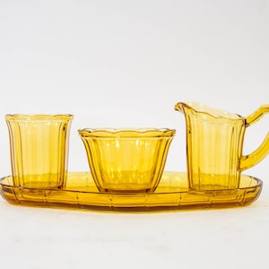 Op de afbeelding: Een set amberkleurig glazen suiker en room met een bijpassend dienblad. De set bestaat uit een kan, een suikerpot en twee kleine kopjes.