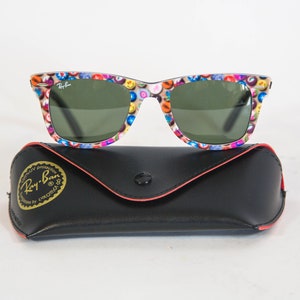 Puede incluir: Gafas de sol Ray-Ban con un patr&oacute;n multicolor de peque&ntilde;os iconos circulares en la montura y lentes de color verde oscuro, sobre un estuche protector negro de piel sint&eacute;tica con el logotipo dorado de Ray-Ban y el texto 100% UV protection.