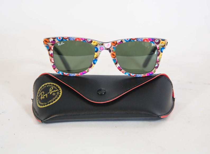 Puede incluir: Gafas de sol Ray-Ban con un patr&oacute;n multicolor de peque&ntilde;os iconos circulares en la montura y lentes de color verde oscuro, sobre un estuche protector negro de piel sint&eacute;tica con el logotipo dorado de Ray-Ban y el texto 100% UV protection.