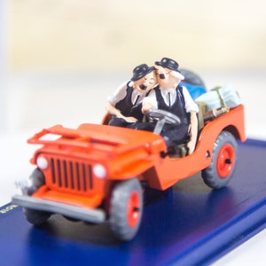May include: A red toy jeep with two figures in the front seats. The figures are wearing black hats and black and white clothing. The jeep has black tires and a black grill. The jeep is on a blue display base.