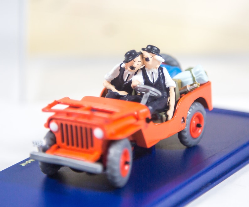 May include: A red toy jeep with two figures in the front seats. The figures are wearing black hats and black and white clothing. The jeep has black tires and a black grill. The jeep is on a blue display base.