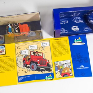 May include: A blue box with a clear plastic window showing a red jeep and a yellow box with a comic book image of a red jeep. The comic book image has the text 'Rron Rron' and 'TINTIN' on it. The blue box has the text 'Tintin' and 'Kuifje en de Zwarte Goud' on it.