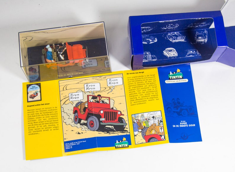 May include: A blue box with a clear plastic window showing a red jeep and a yellow box with a comic book image of a red jeep. The comic book image has the text 'Rron Rron' and 'TINTIN' on it. The blue box has the text 'Tintin' and 'Kuifje en de Zwarte Goud' on it.