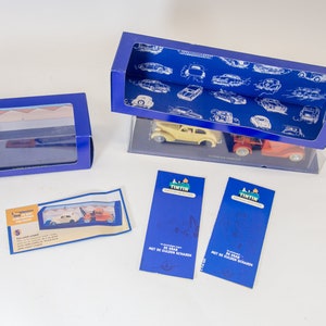 Puede incluir: Dos cajas azules con ventanas de pl&aacute;stico transparente que contienen un coche amarillo y un cami&oacute;n rojo. Las cajas est&aacute;n sobre una superficie blanca. Hay dos tarjetas azules con el texto "Tintin" y "De Zwaar" en ellas.
