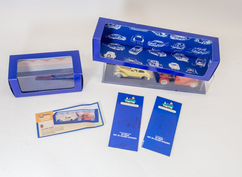 Puede incluir: Dos cajas azules con ventanas de pl&aacute;stico transparente que contienen un coche amarillo y un cami&oacute;n rojo. Las cajas est&aacute;n sobre una superficie blanca. Hay dos tarjetas azules con el texto "Tintin" y "De Zwaar" en ellas.