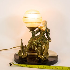 Puede incluir: Una l&aacute;mpara de mesa vintage con una base dorada que presenta una figura femenina sosteniendo una pantalla de l&aacute;mpara en forma de globo con rayas dor&eacute;es. La base tambi&eacute;n presenta dos figuras de cisnes. La l&aacute;mpara est&aacute; sobre una base de m&aacute;rmol negro.