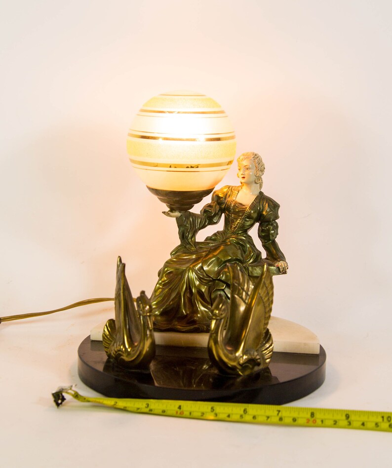 Puede incluir: Una l&aacute;mpara de mesa vintage con una base dorada que presenta una figura femenina sosteniendo una pantalla de l&aacute;mpara en forma de globo con rayas dor&eacute;es. La base tambi&eacute;n presenta dos figuras de cisnes. La l&aacute;mpara est&aacute; sobre una base de m&aacute;rmol negro.