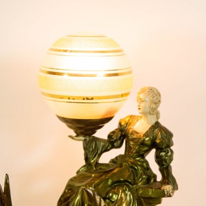 Puede incluir: Una l&aacute;mpara de mesa vintage con una base dorada y una pantalla de globo blanco con rayas doradas. La base presenta una figura esculpida de una mujer con un vestido fluido.