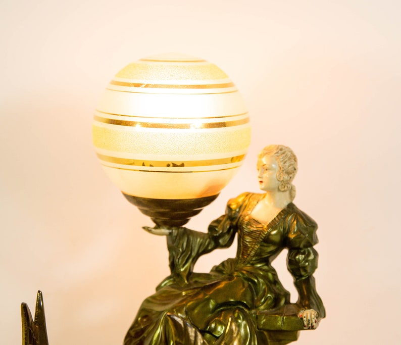 Puede incluir: Una l&aacute;mpara de mesa vintage con una base dorada y una pantalla de globo blanco con rayas doradas. La base presenta una figura esculpida de una mujer con un vestido fluido.