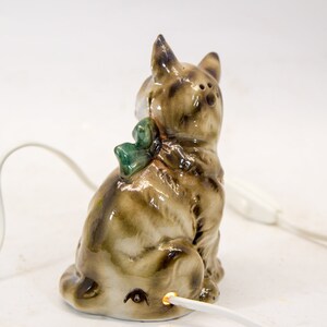 May include: A ceramic cat figurine with a green bow, sitting on a white surface with a white cord plugged into the bottom. The cat is brown and white with a black nose and eyes.