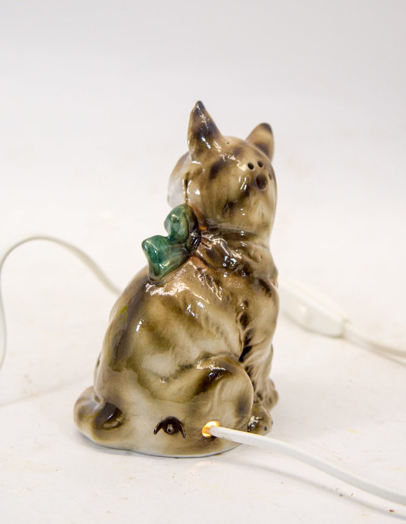 May include: A ceramic cat figurine with a green bow, sitting on a white surface with a white cord plugged into the bottom. The cat is brown and white with a black nose and eyes.