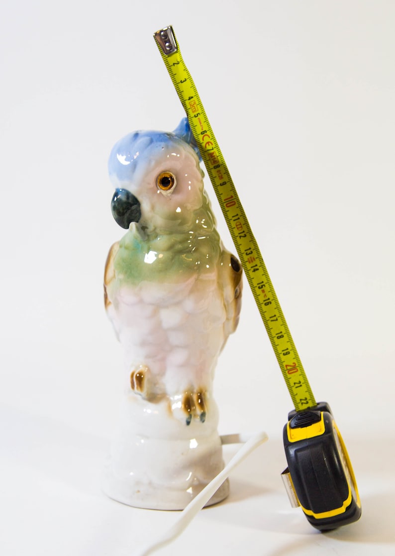 May include: A porcelain parrot figurine with blue, green, and pink painted details, standing on a white base. A yellow measuring tape is next to the figurine, measuring approximately 20 inches.