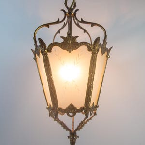 Puede incluir: Lámpara colgante ornamentada de estilo vintage. La lámpara presenta un marco de metal color bronce con intrincados detalles de filigrana y florales. La luz tiene paneles de vidrio esmerilado con un patrón de hojas, y una fuente de luz brillante es visible en el interior.
