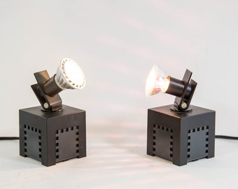 May include: Two black cube-shaped spotlights with adjustable heads. Each spotlight has a dark brown lamp holder and a bright light bulb. The cubes have a square pattern on the sides. The background is white.