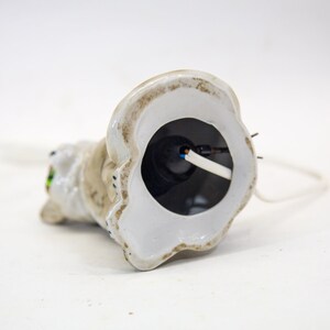 May include: White ceramic cat figurine with green eyes. The base of the figurine is hollow and reveals a white wire connected to a black electrical component.