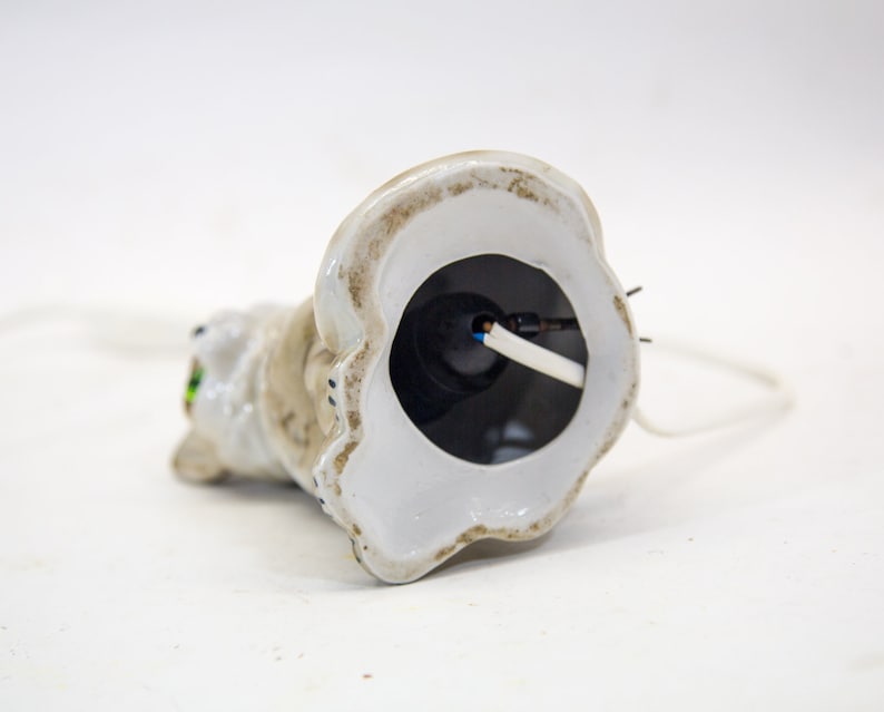 May include: White ceramic cat figurine with green eyes. The base of the figurine is hollow and reveals a white wire connected to a black electrical component.