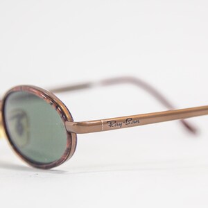 May include: Brown oval-shaped sunglasses with green lenses and a brown metal frame. The sunglasses have the text "Ray-Ban" printed on the frame.