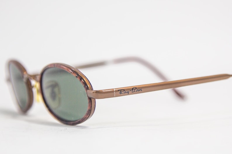 May include: Brown oval-shaped sunglasses with green lenses and a brown metal frame. The sunglasses have the text "Ray-Ban" printed on the frame.