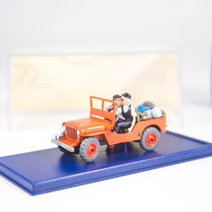 May include: A red toy jeep with two figures inside. The jeep has black wheels and a blue base. The figures are wearing black and white clothing.