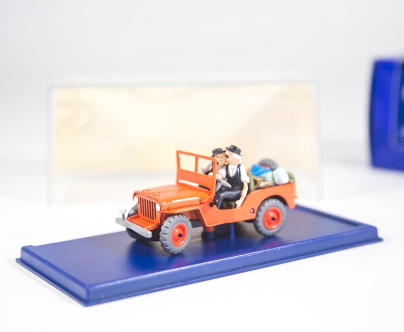 May include: A red toy jeep with two figures inside. The jeep has black wheels and a blue base. The figures are wearing black and white clothing.