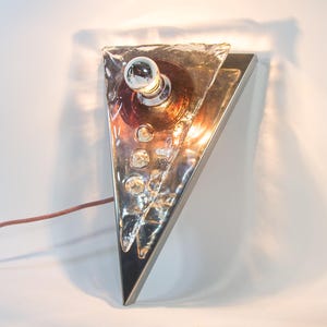 May include: A contemporary wall sconce in a triangular shape, with a silver metal frame and a textured glass shade. It has a small, round light bulb and a brown power cord. The design is geometric and modern.